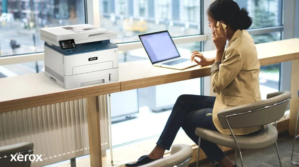 Une personne est assise à une table haute près d'une grande fenêtre, parlant au téléphone tout en travaillant sur un ordinateur portable. Une imprimante multifonction Xerox B235DNI est judicieusement placée sur la table à côté d'elle, suggérant une utilis