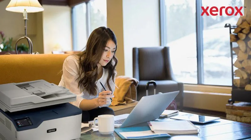 Une femme travaille sur un ordinateur portable dans un bureau ou un salon confortable, avec une imprimante Xerox compacte visible au premier plan, indiquant une intégration transparente de l'impression avec les flux de travail numériques.