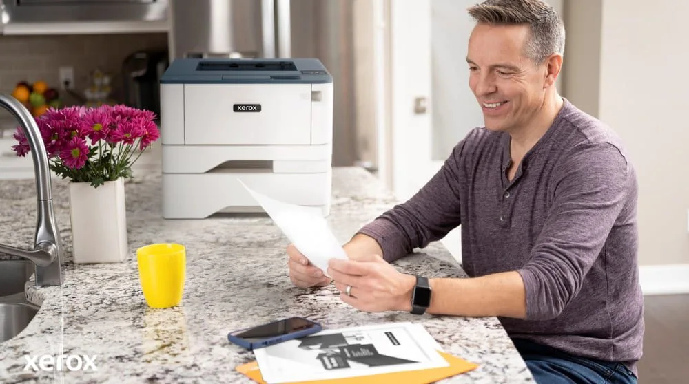 Une personne sourit tout en examinant un document sur un comptoir de cuisine, avec une imprimante Xerox compacte et une tasse jaune vif à proximité, suggérant une impression et une gestion de documents transparentes dans un environnement domestique ou de