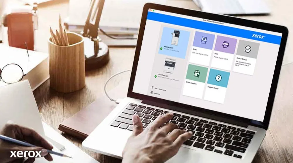 Les mains d'une personne sont posées sur un clavier d'ordinateur portable et l'écran affiche l'interface Xerox Print and Scan Experience avec des options telles que Numériser, Imprimer, État de l'appareil, Commander des fournitures et Centre d'assistance,