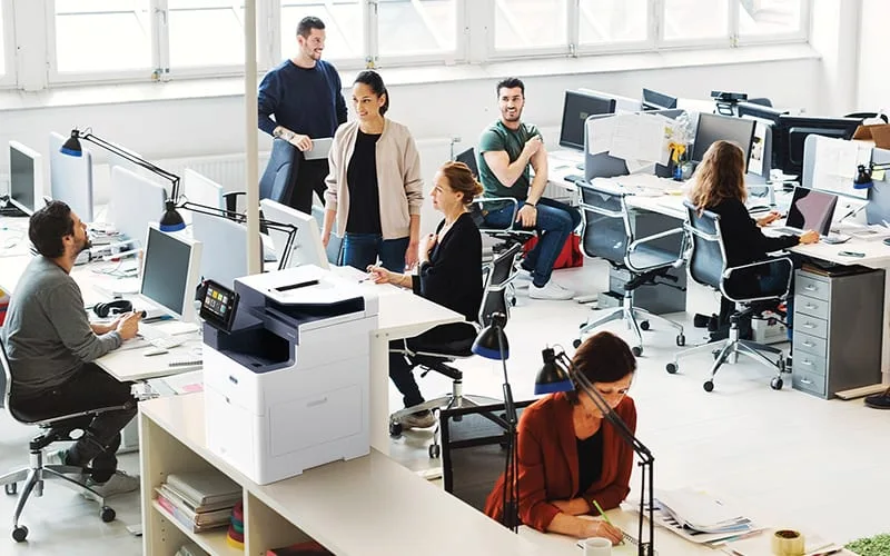 Un groupe de professionnels collaborant dans un bureau, utilisant des ordinateurs et l'imprimante multifonction Xerox® VersaLink® B625 pour leurs tâches.