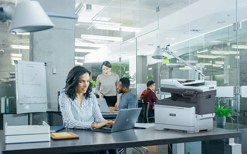 Femme professionnelle tapant sur un ordinateur portable au bureau dans un environnement de bureau à côté d'une imprimante Xerox® C325