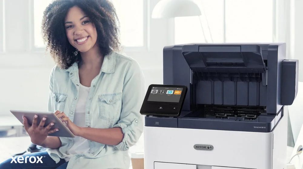 Une personne souriante est assise à côté d'une imprimante Xerox VersaLink B610 blanche, tenant une tablette et regardant la caméra, soulignant la nature conviviale des solutions Xerox.