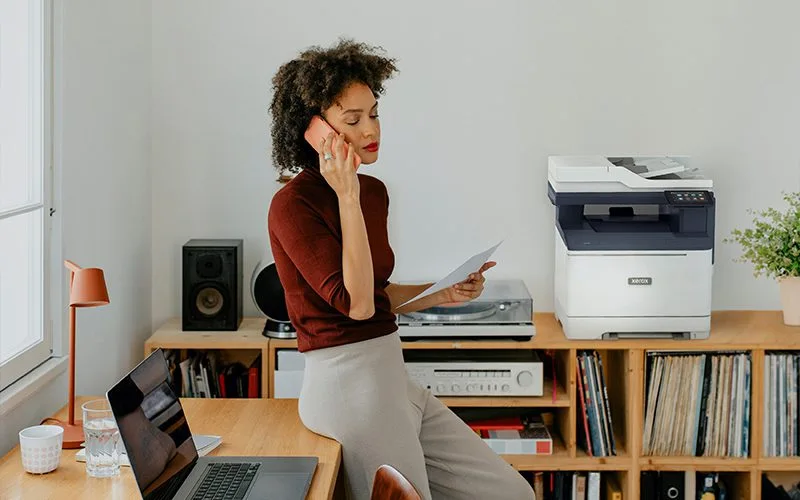 Une femme assise à un bureau en train de passer un appel à côté d'une imprimante Xerox® C325