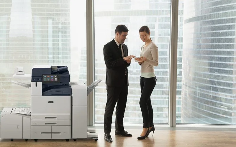Deux personnes en tenue professionnelle dans un bureau discutent de documents à côté d'une imprimante multifonction Xerox® AltaLink® série B8200